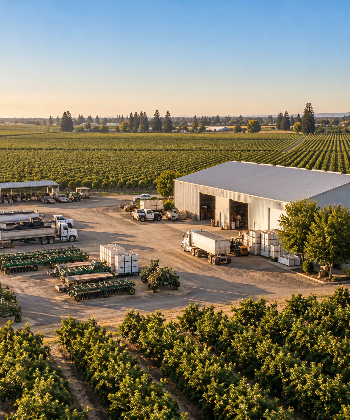 Aerial view of the Ceja Farms yard at golden hour, with trucks, disc harrows, harvest bins and a large equipment barn between orchards